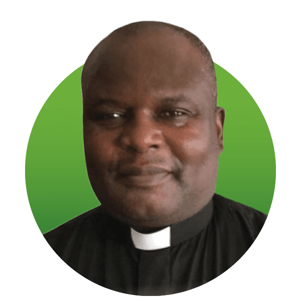 A man wearing a clergy shirt with a white clerical collar stands in front of a green background, looking towards the camera.