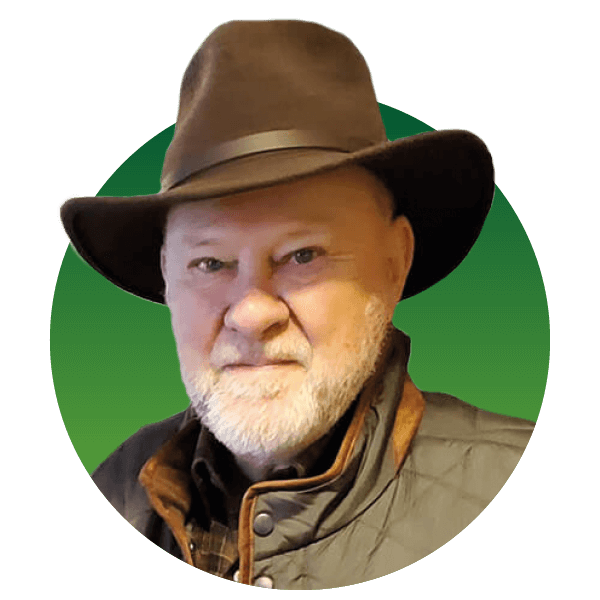 A person with a white beard wearing a brown hat and a dark quilted jacket, against a green background.